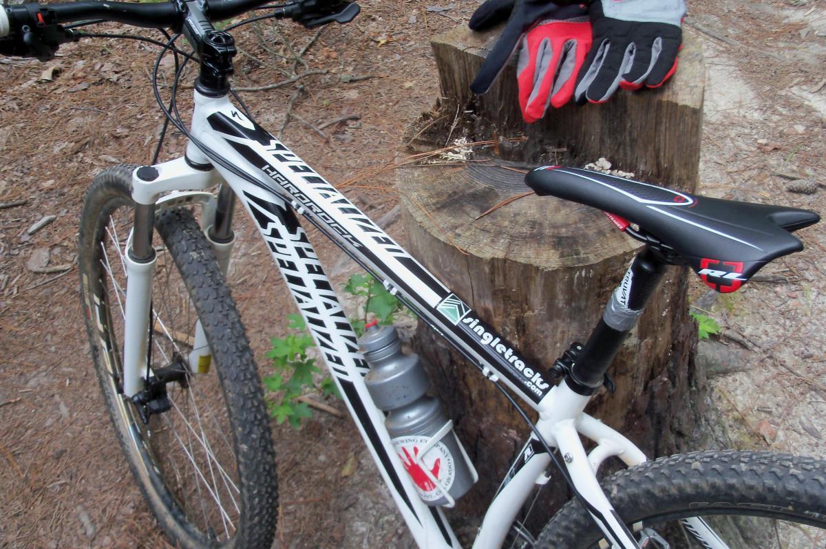 Image of a white mountain bike resting on a forest floor, next to a wooden stump. The bike features a water bottle attached to its frame and red and gray gloves placed on the stump in the background. Pine needles and a few small plants are visible in the surrounding area. Harbison State Forest mountain bike trail.