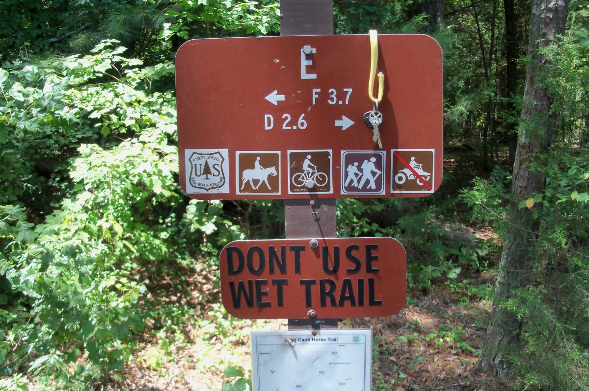 Trail sign indicating directions and distance for various activities, including horseback riding (3.7 miles), biking, and hiking, with a warning not to use the wet trail. The sign is surrounded by greenery in a wooded area. Long Cane Horse Trail mountain bike trail.