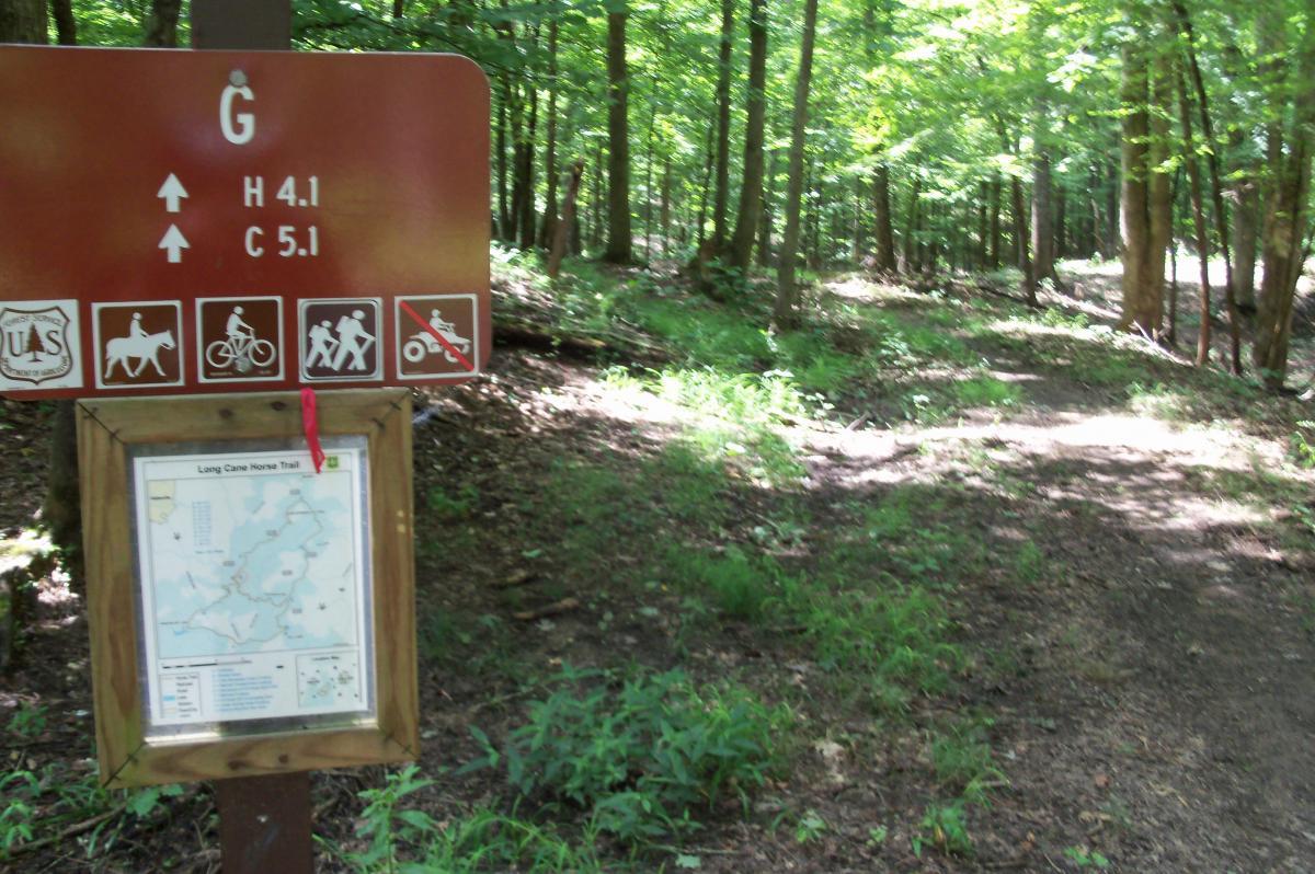 A trail sign indicating the Long Cane Horse Trail in a wooded area. The sign features directional arrows and mileage markers for hiking and cycling. It includes icons for horse riding, biking, and hiking, as well as a prohibition sign for motorized vehicles. A map of the trail is displayed below the sign. Surrounding the sign is lush green foliage and a dirt path leading into the forest. Long Cane Horse Trail mountain bike trail.