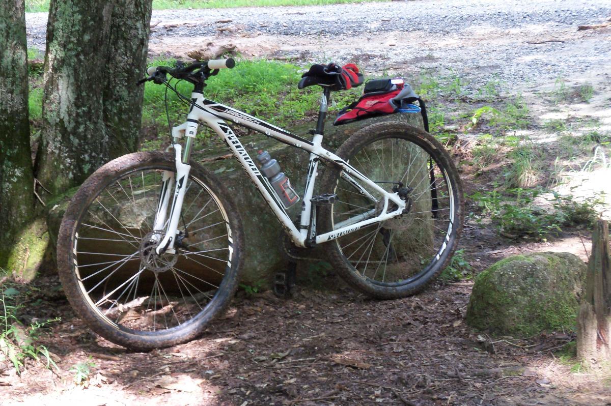A white mountain bike resting against a tree, with a water bottle in the frame. The ground is covered in leaves and dirt, and there are stones and greenery in the background. Long Cane Horse Trail mountain bike trail.