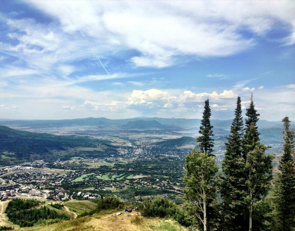 A panoramic view of a lush valley surrounded by mountains under a partly cloudy sky. In the foreground, tall evergreen trees frame the scene, while a small town is visible in the valley below, with green fields and winding roads. The distant mountains add depth to the landscape. Steamboat Ski Resort mountain bike trail.