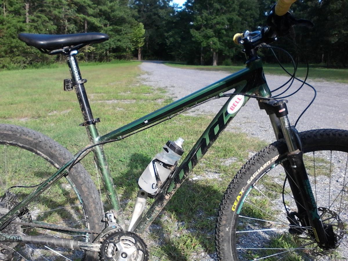 Kona Mahuna: A close-up view of a mountain bike parked on a gravel path surrounded by green grass and trees. The bike features a dark green frame, a black seat, and a water bottle mounted on the frame. The wheels show some dirt, indicating recent use on trails.
