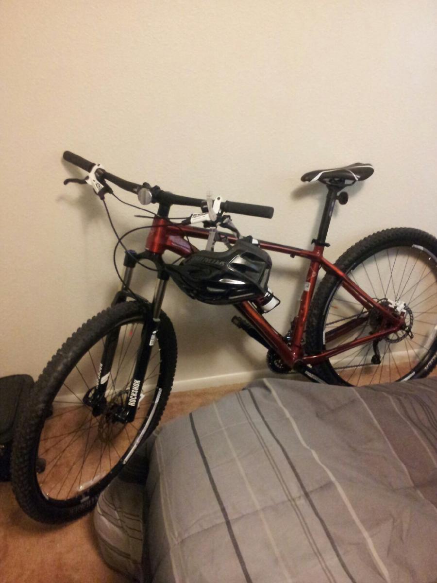 Trek Stache: A red mountain bike leaning against a wall inside a room, with a black helmet attached to the handlebars. The bike is positioned near a bed with gray and white striped bedding and a piece of luggage on the floor.