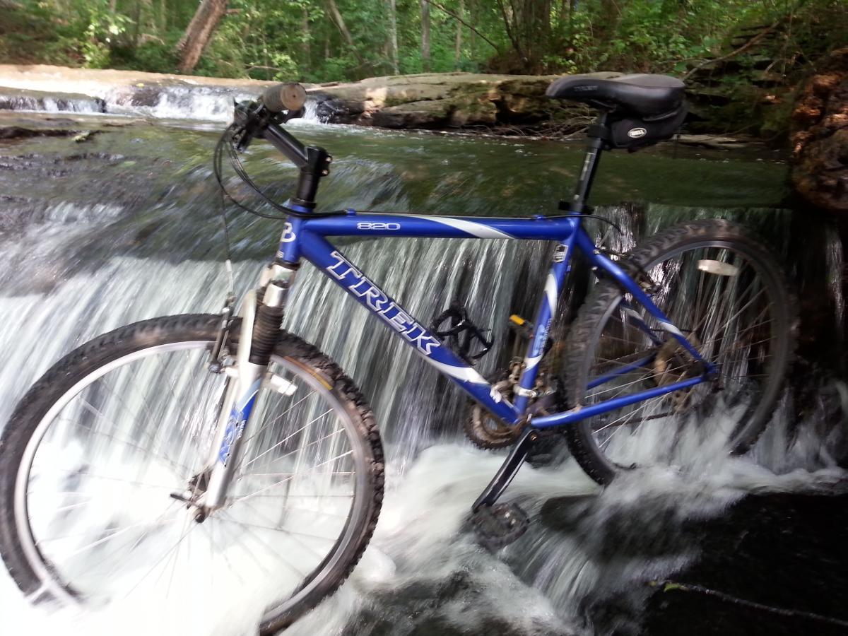 Trek 820: A blue Trek mountain bike positioned on a rocky surface near a flowing stream, with water cascading over the rocks around the bike. Lush greenery can be seen in the background.