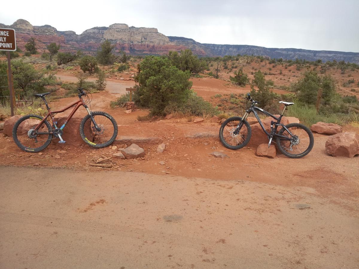 Two mountain bikes are parked on a dirt path surrounded by red rock terrain and sparse vegetation. In the background, there are layered rock formations and a partially cloudy sky. A sign indicating a scenic viewpoint is visible in the left side of the image. Chuck Wagon mountain bike trail.