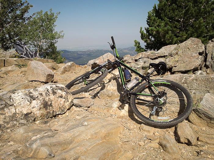A mountain bike resting against rocky terrain, surrounded by trees and mountains in the background. The bike is angled, showcasing its wheels and frame, with a clear sky above. Bergen Peak mountain bike trail.