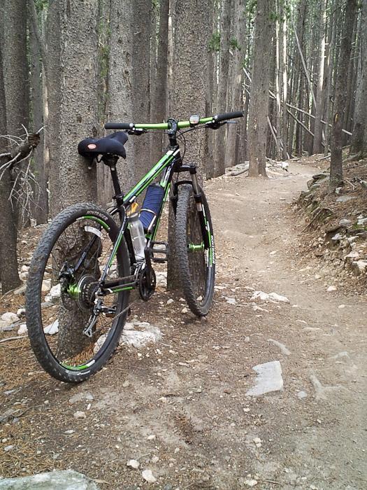 A green and black mountain bike is parked on a dirt trail surrounded by tall trees, with a narrow path winding through the forest in the background. The bike features thick tires and a visible water bottle attached to the frame. Bergen Peak mountain bike trail.
