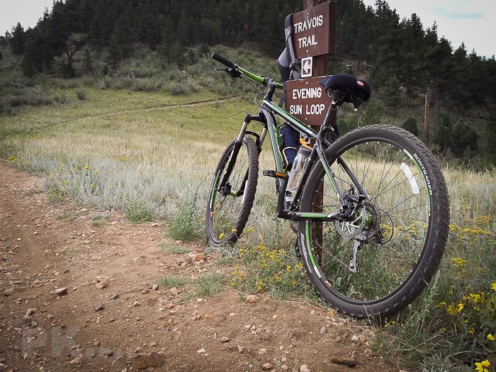 A mountain bike leaning against a trail sign that reads "Travois Trail" and "Evening Sun Loop," with a partially visible grassy path leading into the background, surrounded by trees and wildflowers. Centennial Cone Park mountain bike trail.