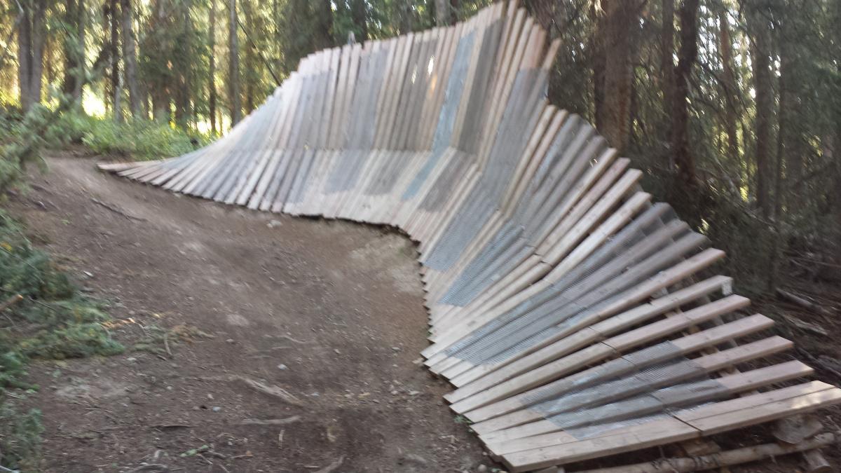 A wooden bike ramp constructed from slats, curving gently along a forested trail, surrounded by tall trees and greenery. The ramp features a smooth incline designed for mountain biking, blending into the natural landscape. Evolution Bike Park at Crested Butte Mountain Resort mountain bike trail.