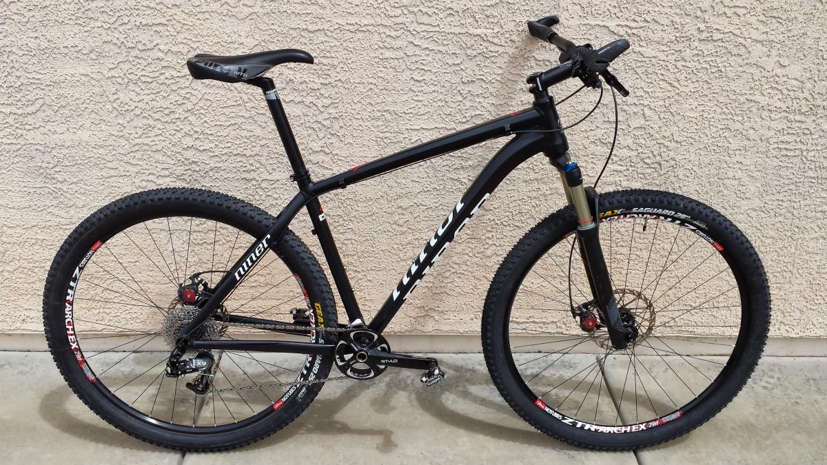 Niner EMD 9: A black mountain bike with thick tires and a lightweight frame is positioned against a textured beige wall. The bike features a front suspension fork, a curved handlebar, and a sleek saddle, showcasing a modern design for outdoor cycling.