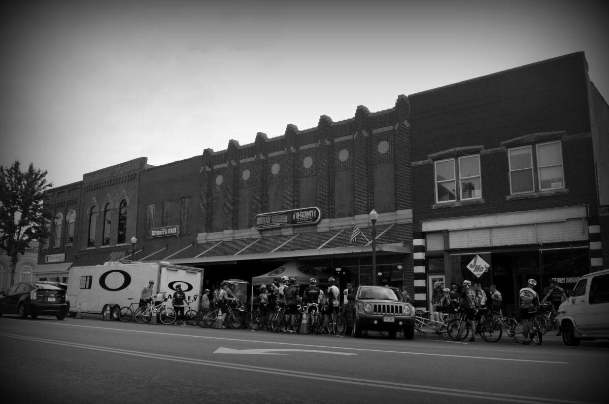 Black and white image of a historic brick storefront with multiple windows and a sloped roof. Outside, a gathering of cyclists with their bikes is visible next to a trailer featuring an "O" logo. Cars are parked along the street, and several shop signs are displayed on the building. The scene captures an outdoor cycling event in a small-town setting.