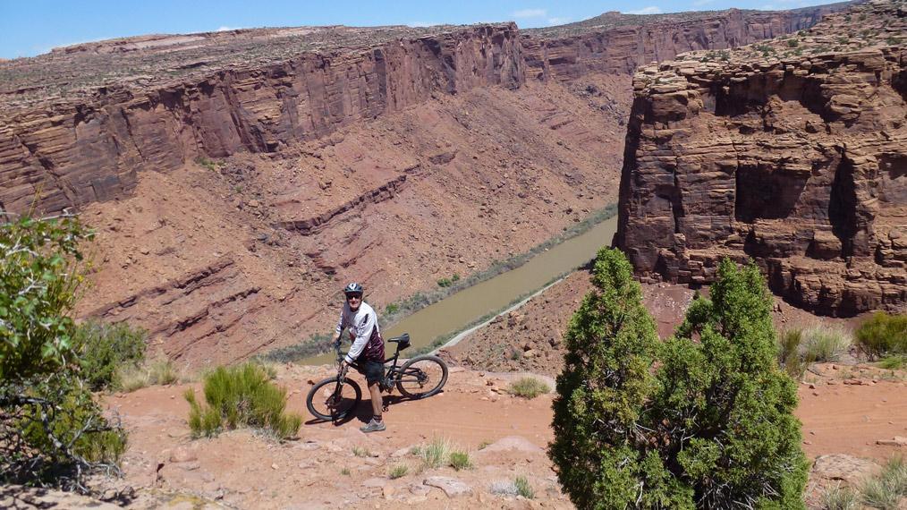 Mountain biker standing beside a bicycle on a rocky path overlooking a canyon with steep cliffs and a river running through the valley below. Lush green shrubs and plants are visible in the foreground against a clear blue sky. Porcupine Rim mountain bike trail.