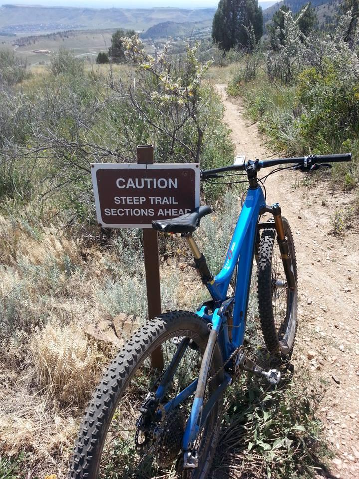 A blue mountain bike parked next to a sign that reads "Caution: Steep Trail Sections Ahead," with a mountainous landscape visible in the background and greenery lining the trail. White Ranch mountain bike trail.