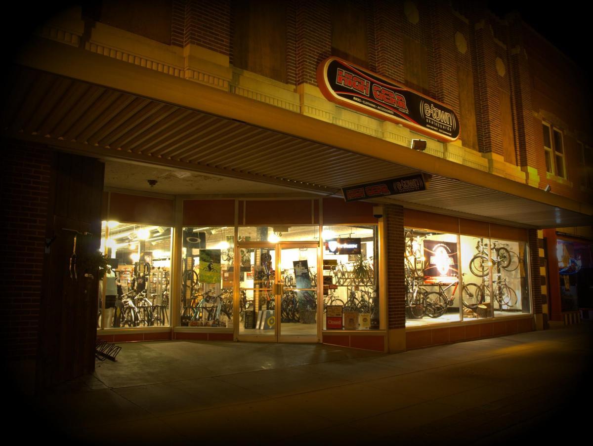 Image of a bike shop storefront at night, featuring large windows displaying various bicycles and cycling gear. The sign above the entrance reads "High Gear e-town." The interior is lit, showcasing a range of bikes and accessories, while the sidewalk in front is empty.