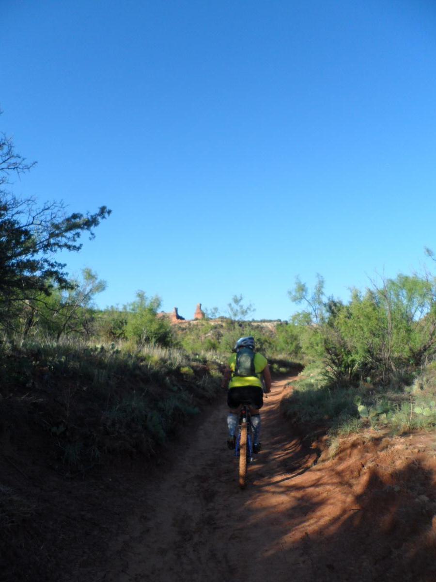 A cyclist riding on a dirt trail surrounded by green vegetation, with a clear blue sky overhead and rock formations visible in the distance. Palo Duro Canyon mountain bike trail.