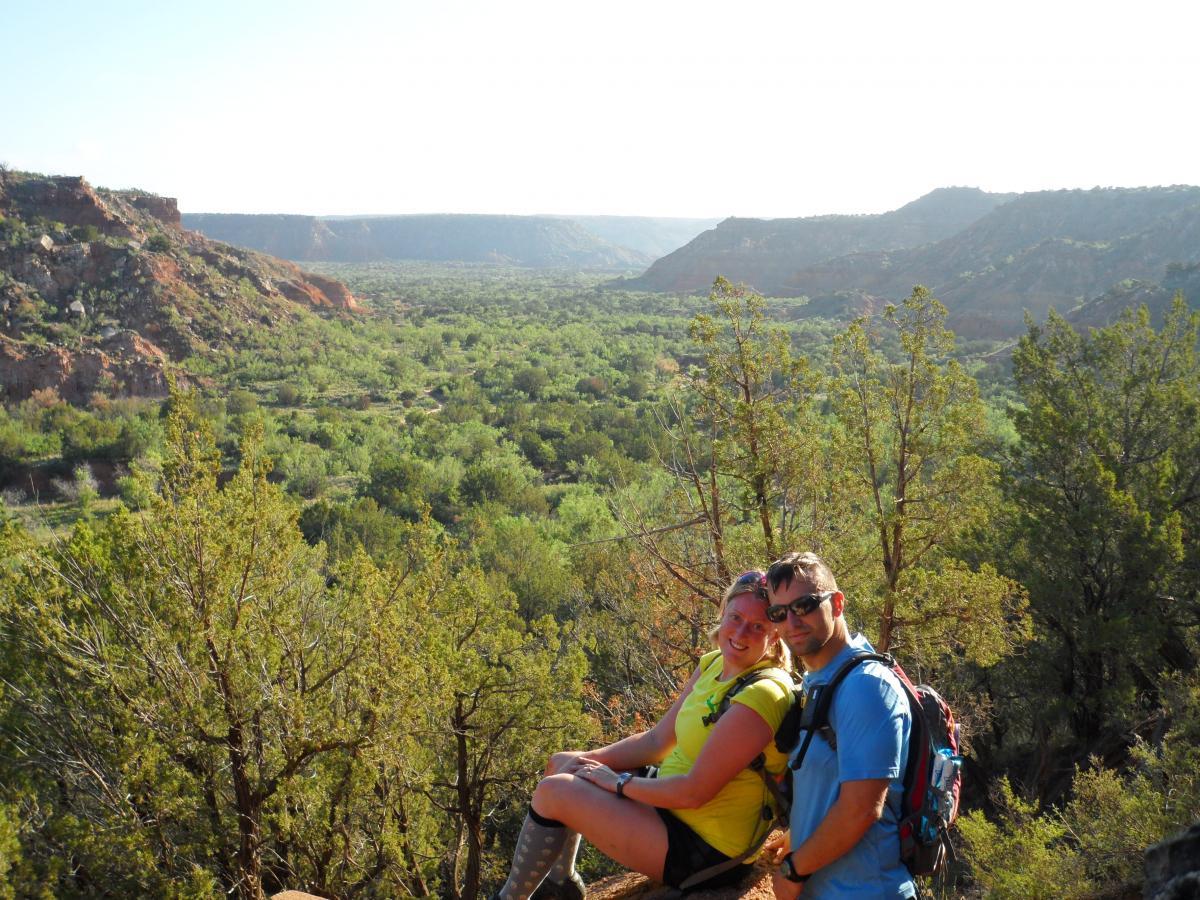 A couple sitting on a rocky ledge, smiling together while overlooking a lush green valley surrounded by red cliffs. The sun is shining brightly, and the landscape features a variety of trees and vegetation. The woman is wearing a bright yellow shirt and shorts, while the man is dressed in a blue shirt with a backpack. Palo Duro Canyon mountain bike trail.