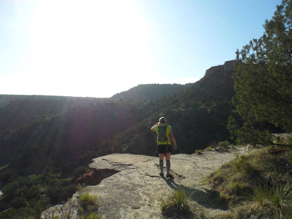 A hiker with a backpack walks along the edge of a rocky cliff, surrounded by rolling green hills and a bright, sunlit sky. The scene captures the beauty of nature and the sense of adventure in outdoor exploration. Palo Duro Canyon mountain bike trail.