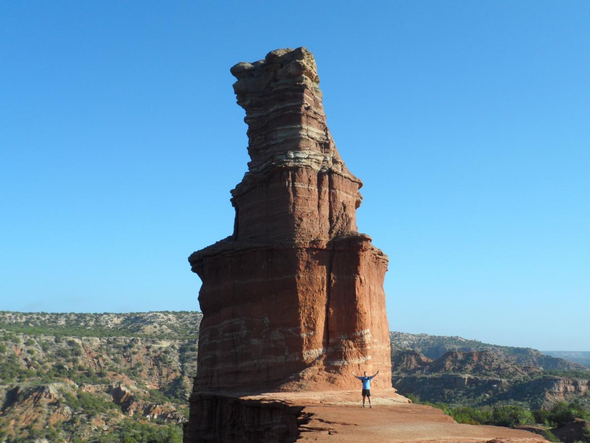 A person standing with outstretched arms at the base of a towering rock formation, known as a monument, against a clear blue sky. The landscape features layered rock formations and rolling hills in the background, highlighting the natural beauty of the area. Palo Duro Canyon mountain bike trail.