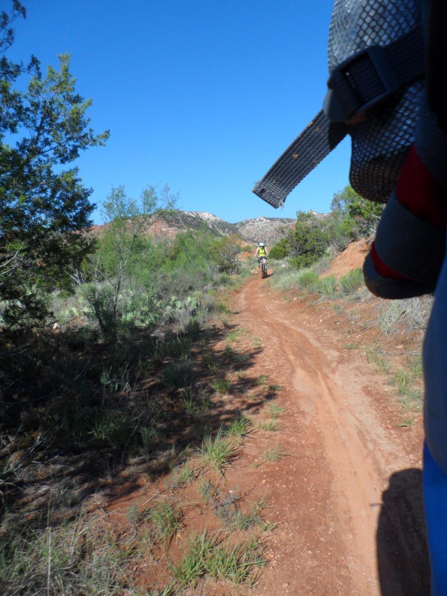 A dirt path winding through a scenic, green landscape with a cyclist riding in the distance. The foreground shows part of a person wearing a harness or backpack, while the background features shrubs, trees, and rocky hills under a clear blue sky. Palo Duro Canyon mountain bike trail.