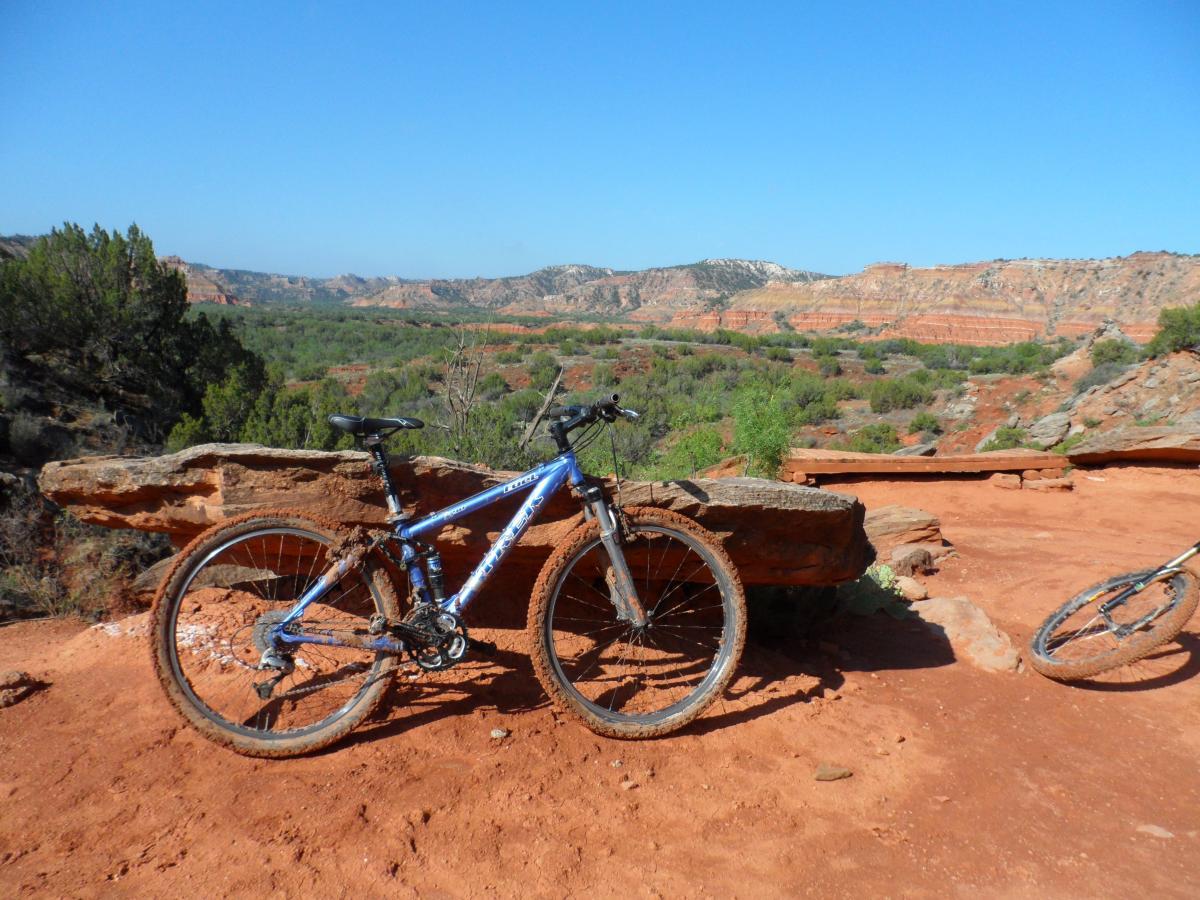 Mountain bikes resting on a rocky outcrop overlooking a scenic canyon landscape filled with red soil, greenery, and layered rock formations under a clear blue sky. Palo Duro Canyon mountain bike trail.