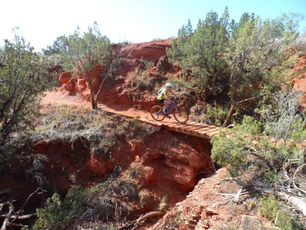 A mountain biker navigates a narrow wooden bridge over a rocky, red terrain, surrounded by small shrubs and trees on a sunny day. Palo Duro Canyon mountain bike trail.