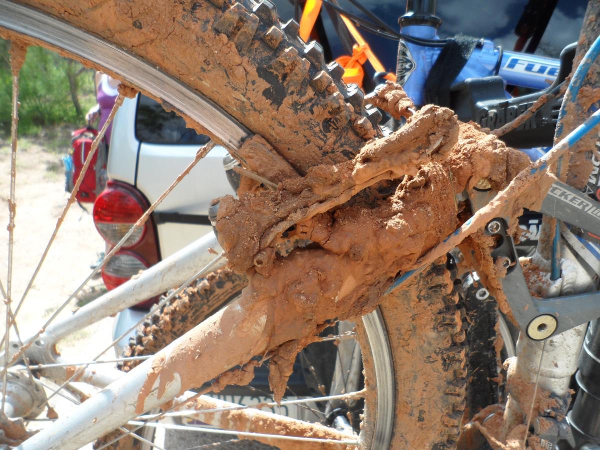 Close-up of a muddy mountain bike rear wheel and drivetrain, showing thick, clumpy mud accumulation on the frame and gears. A blurred vehicle is visible in the background, along with greenery that suggests an outdoor biking environment. Palo Duro Canyon mountain bike trail.