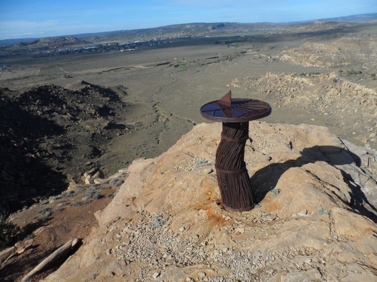 A metal sculpture resembling a twisted pillar stands on a rocky outcrop, overlooking a vast, arid landscape with distant hills and a small town in the background under a blue sky. High Desert Trail System mountain bike trail.