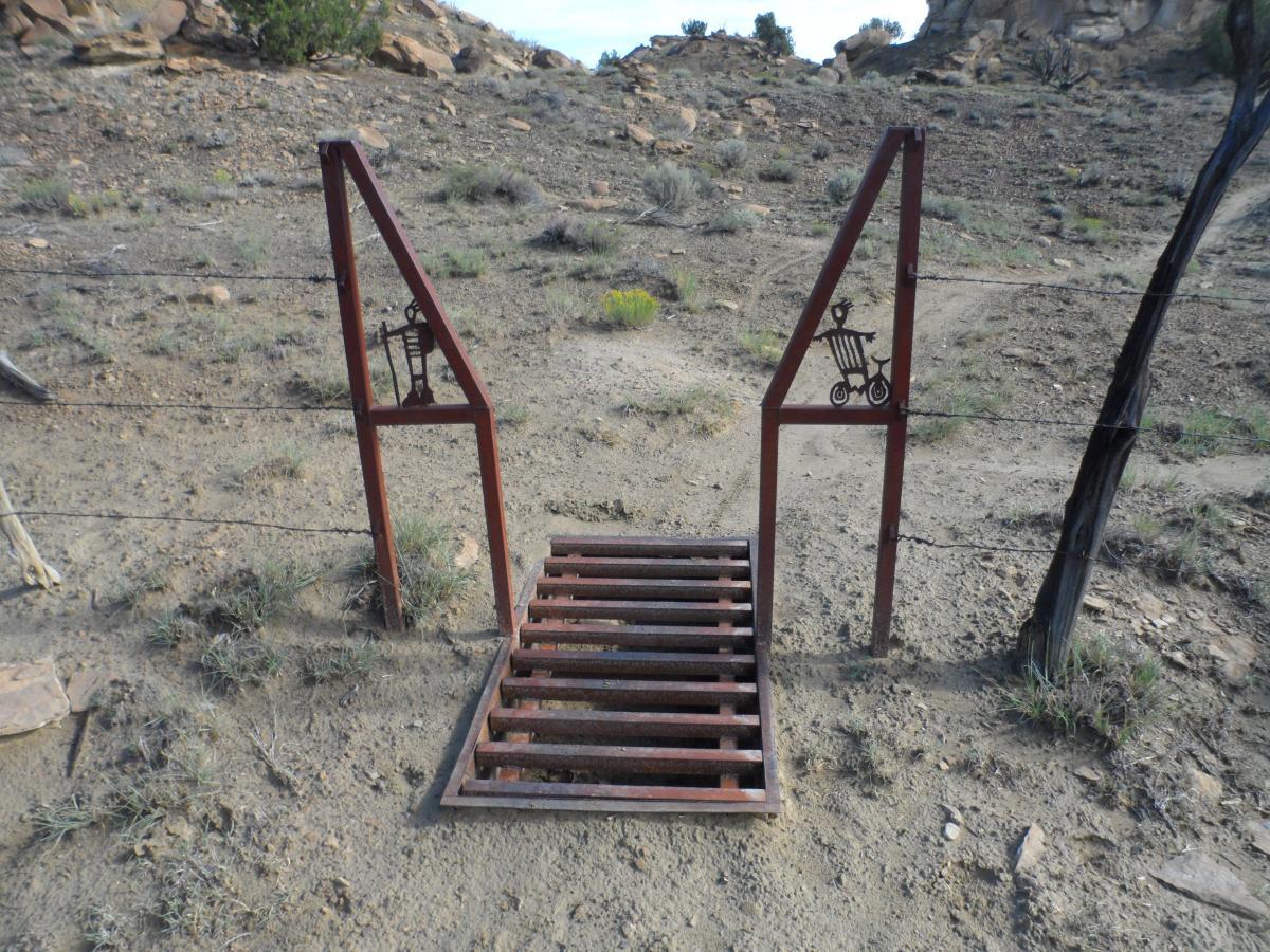 Alt text: A metal cattle guard with artistic designs of a bicycle on one side and a beer mug on the other, positioned on a dirt path surrounded by sparse vegetation and rocky terrain. High Desert Trail System mountain bike trail.