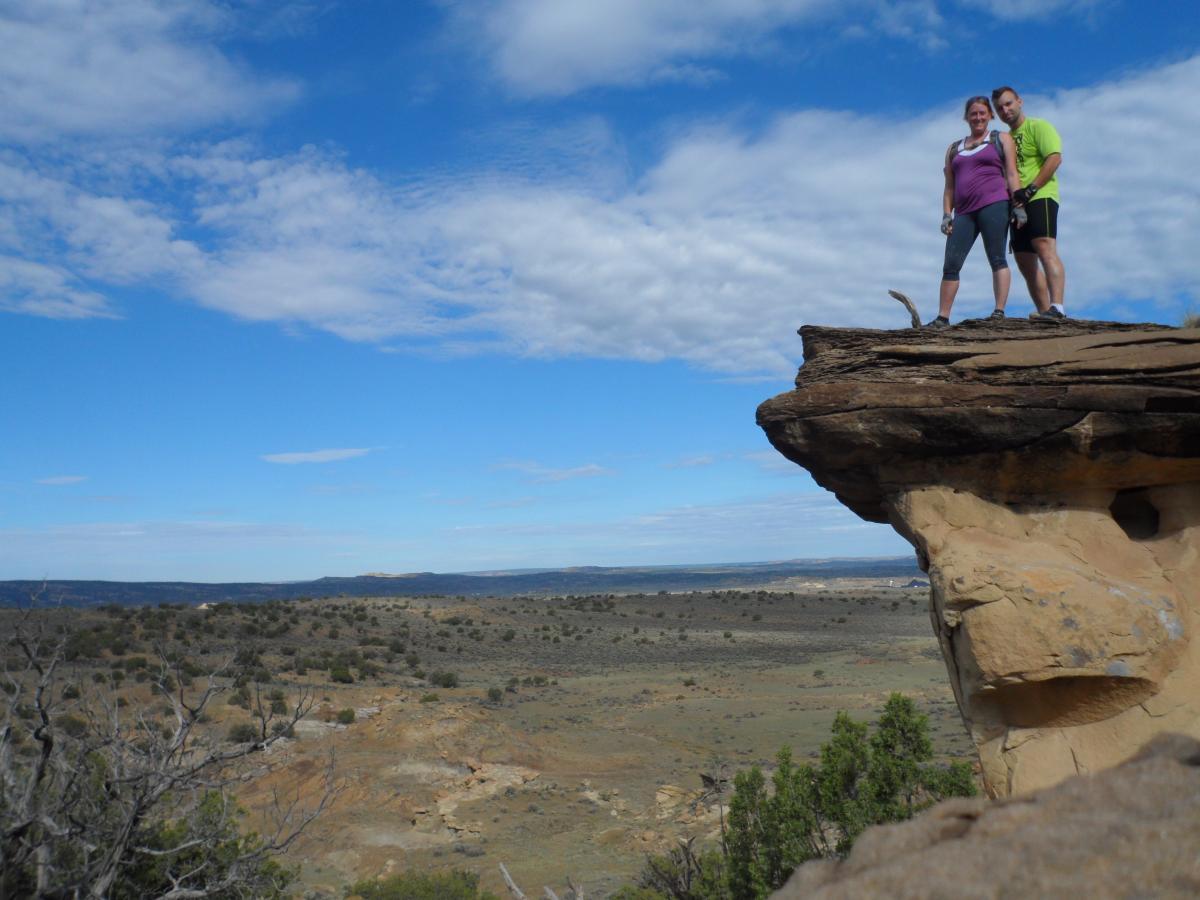 A man and a woman stand on the edge of a rocky outcrop, looking out over a vast, open landscape under a bright blue sky with clouds. The terrain features small shrubs and distant hills, creating a scenic view. The individuals are dressed in athletic clothing, suggesting an outdoor adventure. High Desert Trail System mountain bike trail.