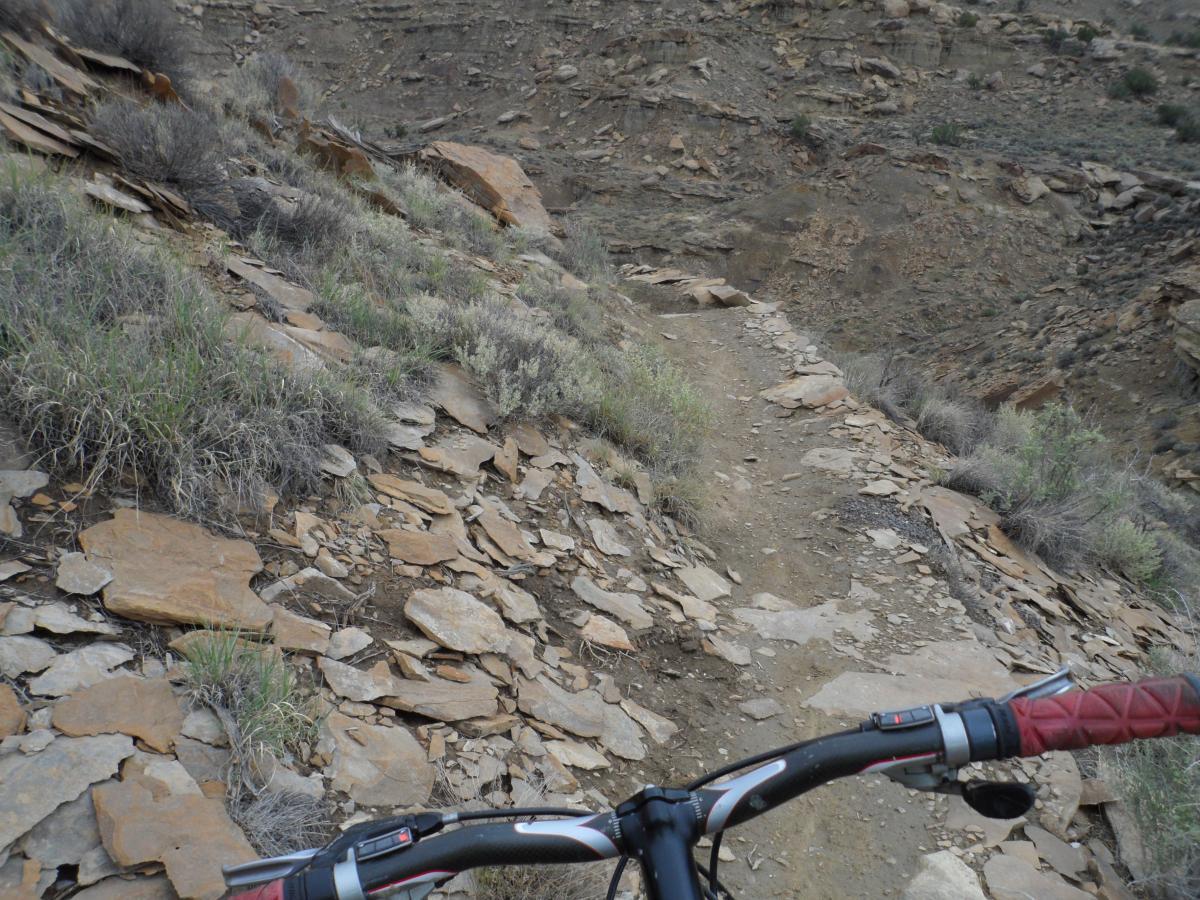 View of a rocky mountain biking trail from the perspective of the bike's handlebars, featuring uneven terrain composed of stone and sparse grass on the slopes, with a steep drop-off visible on the side. High Desert Trail System mountain bike trail.