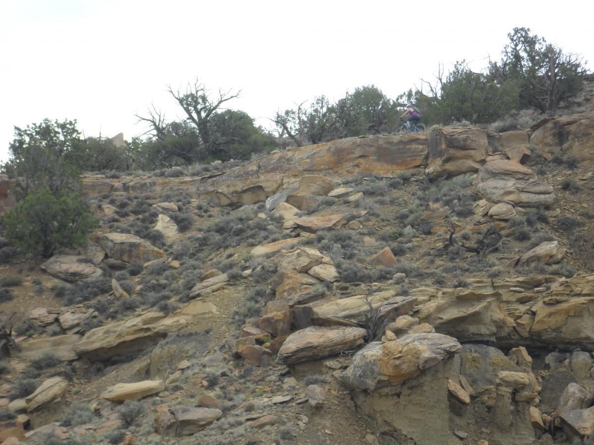 A mountainous landscape featuring rocky terrain and sparse vegetation. In the background, a person can be seen riding a bicycle along a narrow path on the hillside. The sky is overcast, adding a muted light to the scene. High Desert Trail System mountain bike trail.