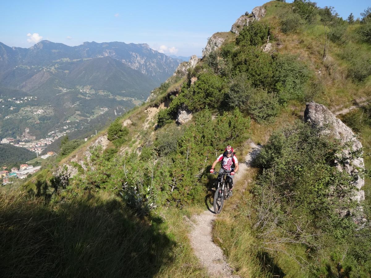 A mountain biker navigating a narrow trail on a hillside, surrounded by lush greenery and rocky formations, with a scenic view of mountains and a valley in the background under a clear blue sky. Valle Cumina e Valcava mountain bike trail.