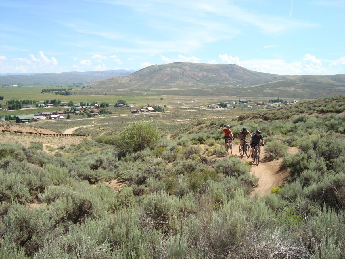 Three mountain bikers navigate a dirt trail through sagebrush-covered hills, with a rural town and green fields visible in the background. The scene is set against a clear blue sky and rolling hills. Hartman Rocks mountain bike trail.