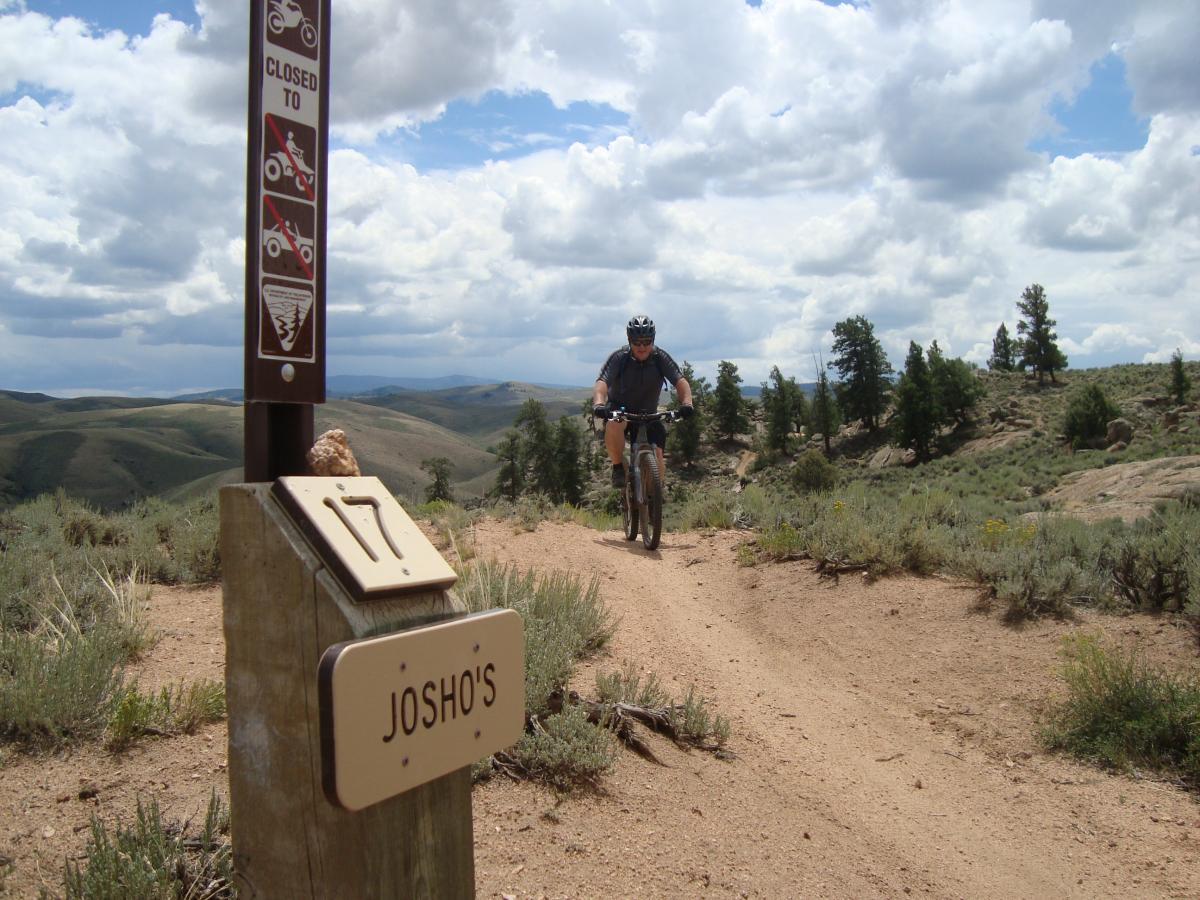 A mountain biker riding on a dirt trail through a mountainous landscape. In the foreground, a wooden trail sign labeled "Josh's" is visible, along with a sign indicating areas that are closed to specific vehicles. The skies are partly cloudy, and the scenery features rolling hills and scattered trees. Hartman Rocks mountain bike trail.