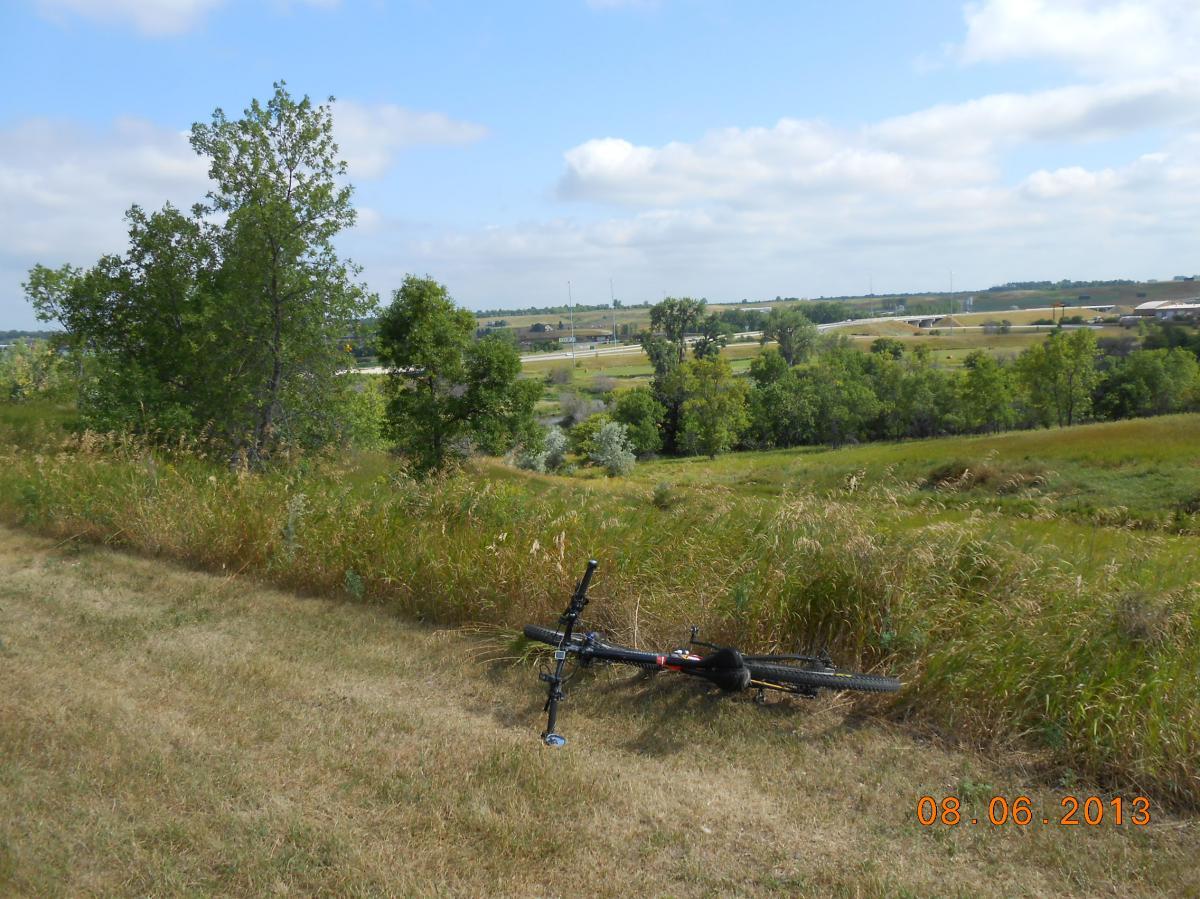 A black bicycle lying on its side in a grassy area, surrounded by tall grasses and trees. In the background, a scenic view of fields and a distant roadway under a partly cloudy sky. The image reflects a serene outdoor setting. White Cloud Nature Trail mountain bike trail.