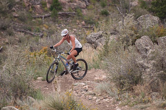 A person riding a mountain bike on a rocky trail surrounded by shrubs and small plants, with a hilly background. The cyclist is focused, wearing a helmet and athletic clothing. Centennial Cone Park mountain bike trail.