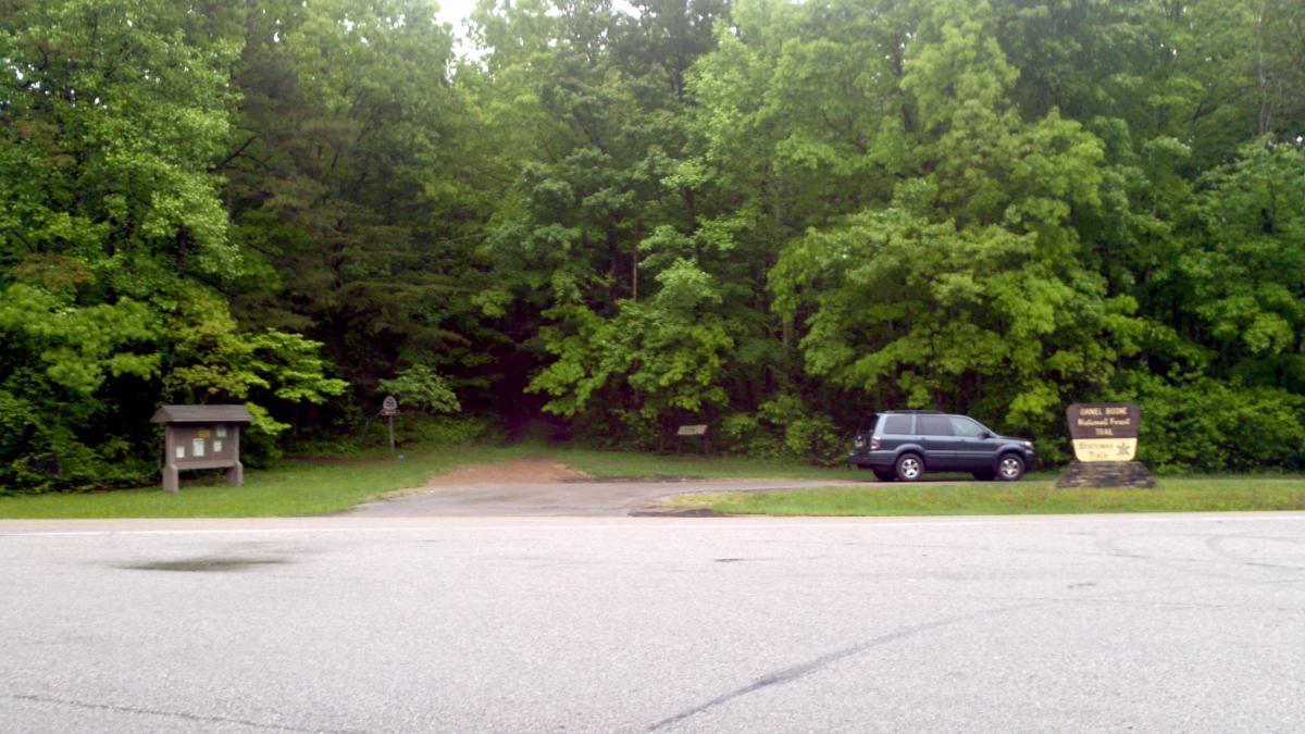 Image showing a gravel parking area at the entrance to a wooded trail. A parked vehicle is visible beside a sign for the Daniel Boone National Forest Trail, along with an information board. The surrounding area is lush with green trees, indicating a natural and inviting outdoor environment. Sheltowee Trace - Laurel Lake Trail mountain bike trail.