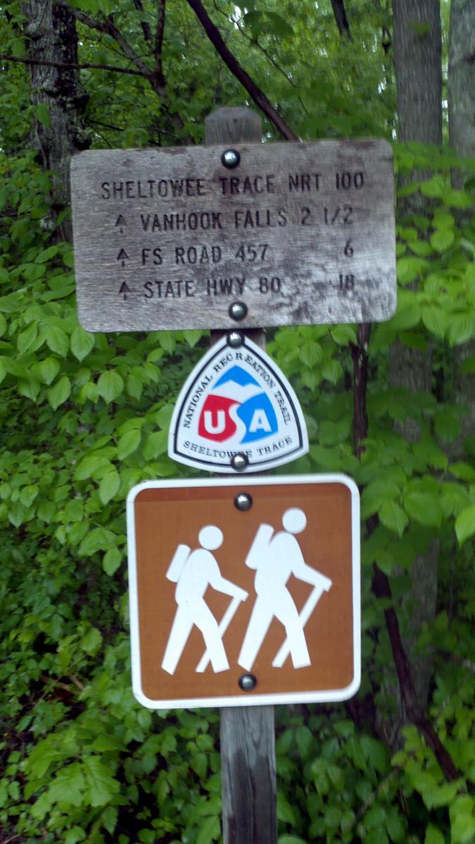 A wooden trail sign indicating directions and distances to various landmarks on the Sheltowee Trace National Recreation Trail, surrounded by green foliage. The sign displays distances to Vanhook Falls (2.5 miles), FS Road 457 (6 miles), and State Highway 80 (18 miles). A blue, red, and white National Recreation Trail emblem and a brown hiking trail marker with two hikers are also visible. Sheltowee Trace - Laurel Lake Trail mountain bike trail.