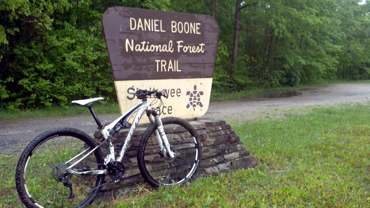 A mountain bike resting against a sign that reads "Daniel Boone National Forest Trail," situated near a gravel path surrounded by green trees and grass. Sheltowee Trace - Laurel Lake Trail mountain bike trail.