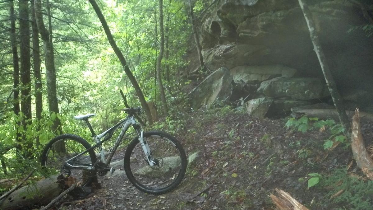 A mountain bike resting on a dirt trail surrounded by lush greenery and trees, with a large rocky outcrop in the background. The scene conveys a peaceful outdoor setting for cycling or hiking. Sheltowee Trace - Laurel Lake Trail mountain bike trail.