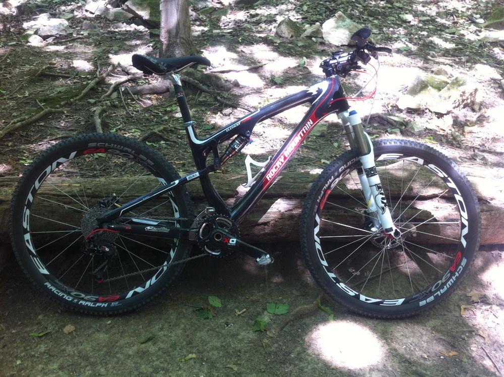 Rocky Mountain Element Team: A mountain bike leaning against a log in a forested area, with sunlight filtering through the trees. The bike features a black and red frame, large wheels with bold white branding, and is equipped with front suspension. Surrounding the bike are green leaves and natural terrain, indicating a suitable environment for outdoor cycling.
