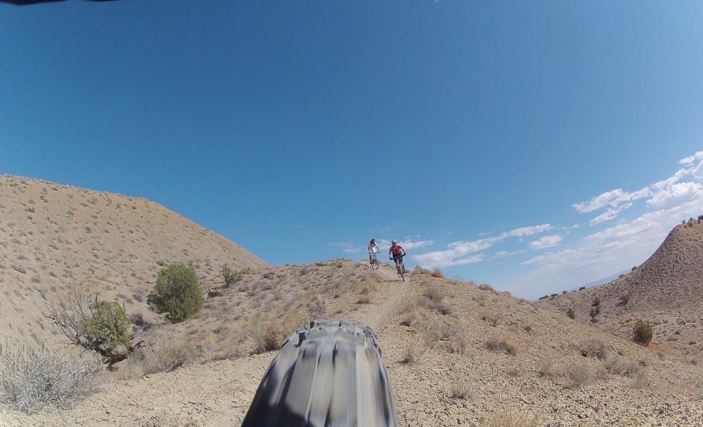 A view of a rugged mountain biking trail under a clear blue sky, featuring two bikers riding uphill on a dirt path surrounded by dry hills and sparse vegetation. The foreground shows a close-up of a mountain bike tire. Zippety Do Dah mountain bike trail.