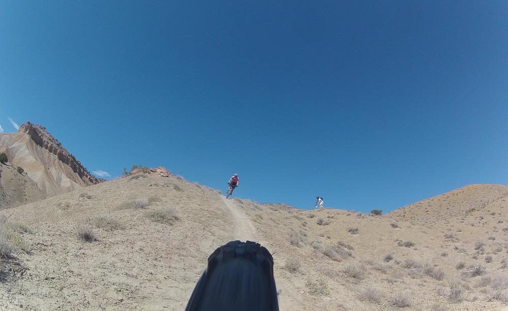 A mountain biking trail captured from a low angle, showing the rear wheel of a bike in the foreground. In the background, a rider in red pedals up a dusty slope against a clear blue sky, with another cyclist visible further up the trail. The terrain is arid and hilly, with scattered vegetation. Zippety Do Dah mountain bike trail.