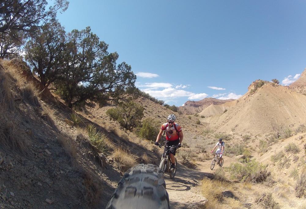 Two mountain bikers riding on a dirt trail through a rocky, arid landscape with hills in the background and clear blue skies above. Zippety Do Dah mountain bike trail.