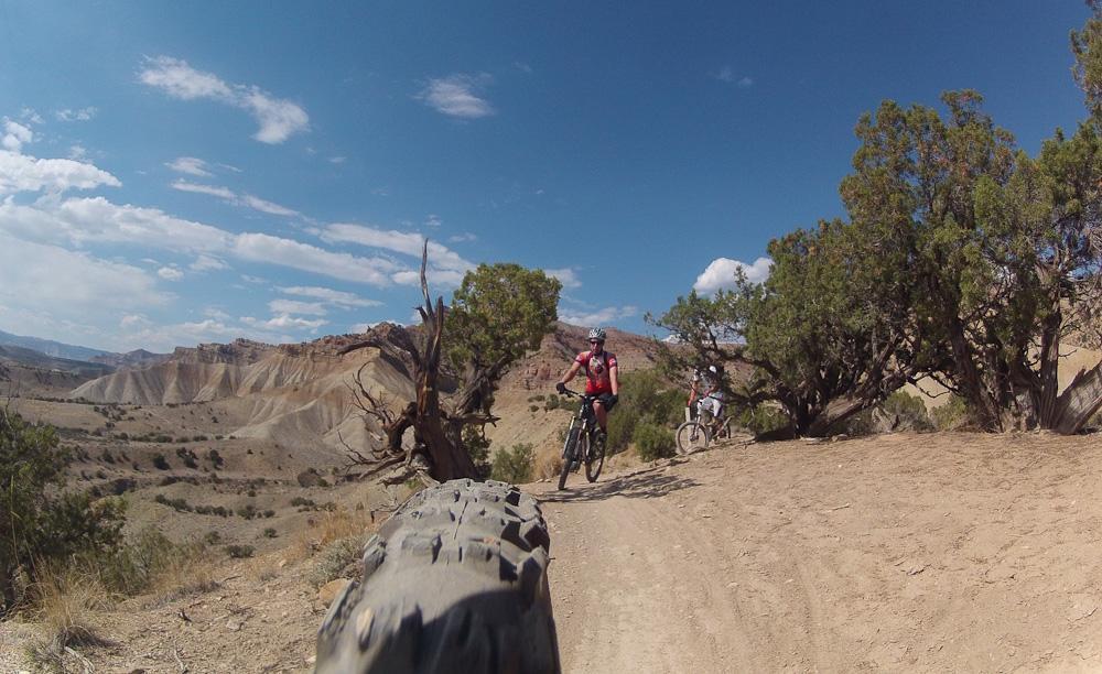 Two mountain bikers riding along a dusty trail surrounded by a scenic landscape of hills and trees under a blue sky with scattered clouds. Zippety Do Dah mountain bike trail.