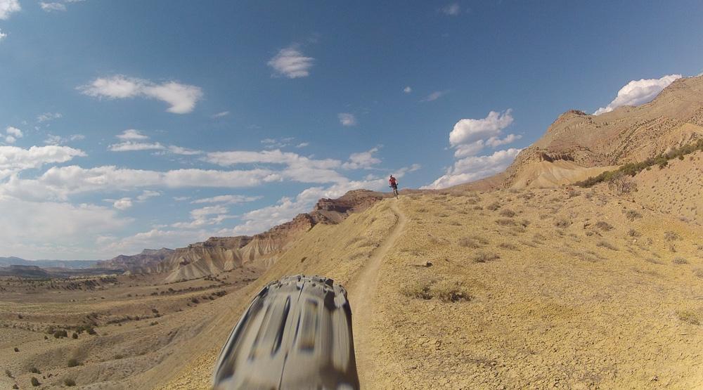 A mountain biking view of a rugged landscape, featuring a dirt trail winding through arid terrain with rocky hills in the background. A cyclist is seen in the distance, riding along the path under a partly cloudy blue sky. Zippety Do Dah mountain bike trail.