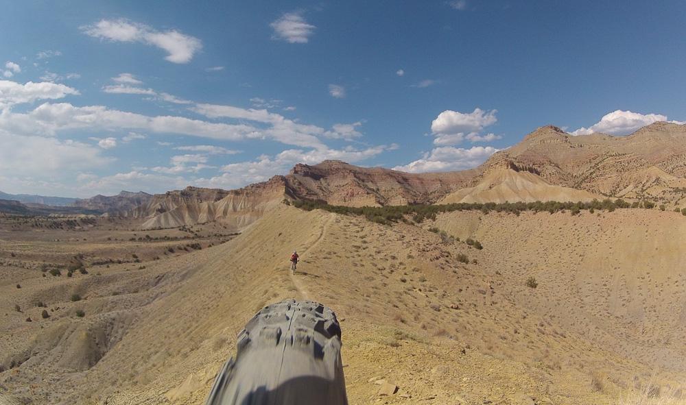 A mountain biker rides along a narrow dirt trail on a hillside, surrounded by rugged terrain and colorful rock formations under a bright blue sky with scattered clouds. Zippety Do Dah mountain bike trail.