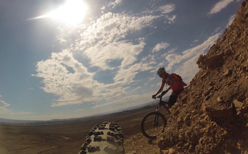 A mountain biker navigating a rocky trail under a bright sky, with the sun shining above and expansive landscapes in the background. The focus is on the cyclist, who is wearing a helmet and riding gear, as they maneuver along a steep incline. Zippety Do Dah mountain bike trail.
