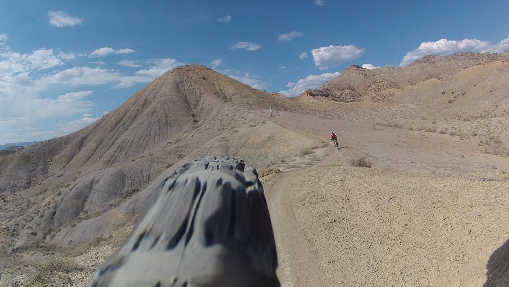 A dirt trail winding through a rugged, arid landscape with two hikers in the distance, surrounded by hills and blue skies dotted with clouds. The foreground shows part of a mountain bike wheel, emphasizing an outdoor adventure setting. Zippety Do Dah mountain bike trail.