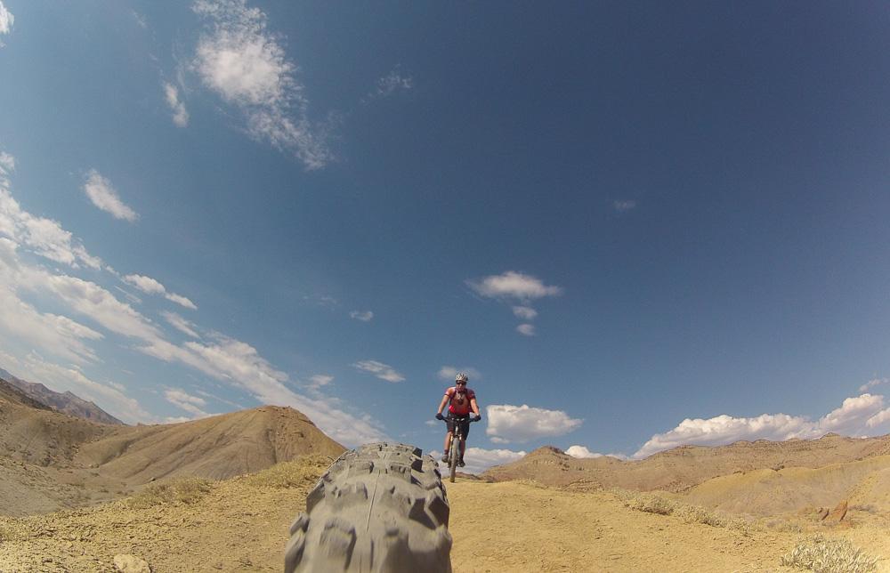 A mountain biker riding along a dirt trail in a rugged landscape, with mountains in the background and a bright blue sky dotted with clouds. The view captures the tire of the bike in the foreground as the rider pedals forward. Zippety Do Dah mountain bike trail.